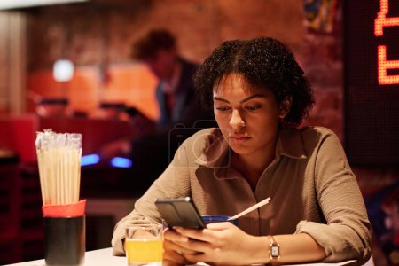 Foto de Joven mujer de negocios seria mirando a través de la información en línea en la pantalla del teléfono inteligente mientras está sentado en la cafetería japonesa y almorzando - Imagen libre de derechos