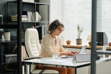 Téléchargez les photos : Vue latérale de l'employeur féminin mature tapant sur le clavier d'ordinateur portable tout en étant assis par le lieu de travail dans le bureau et en travaillant avec des données en ligne - en image libre de droit