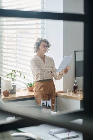 Foto de Mujer experimentada directora ejecutiva mirando a través de documentos financieros o contrato de negocios mientras está de pie por la ventana en el cargo - Imagen libre de derechos