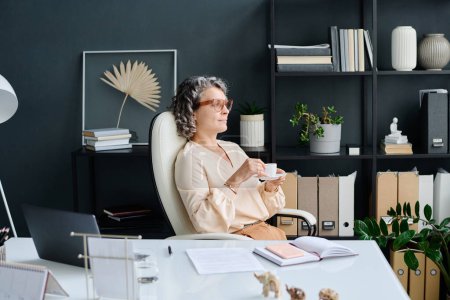 Foto de Ceo femenino maduro reparador con taza de café sentado por el lugar de trabajo en la oficina, mirando a través de la ventana y pensando en algo agradable - Imagen libre de derechos