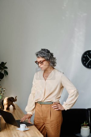 Téléchargez les photos : Femme d'affaires mature avec cheveux gris touches touchantes de clavier d'ordinateur tout en se tenant près du bureau en bois avec une tasse de café - en image libre de droit