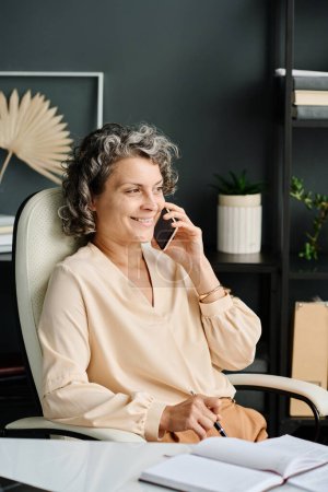 Téléchargez les photos : Femme souriante mature en chemisier beige parlant sur un téléphone portable tout en étant assise dans un fauteuil en cuir blanc confortable près de son lieu de travail - en image libre de droit
