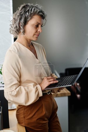 Téléchargez les photos : Analyste féminine mature sérieuse dans des vêtements de luxe silencieux regardant l'écran de l'ordinateur portable et travaillant dans le net ou organisant le travail tout en restant au bureau - en image libre de droit