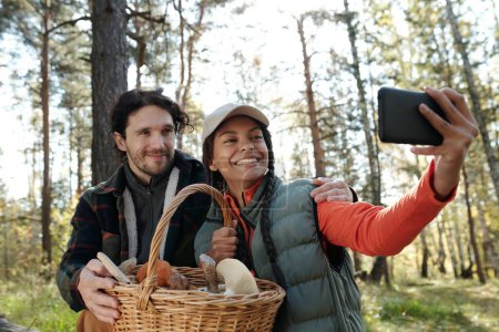 Téléchargez les photos : Jeunes dates interculturelles avec panier de champignons regardant la caméra smartphone avec des sourires pendant selfie dans la forêt d'automne - en image libre de droit