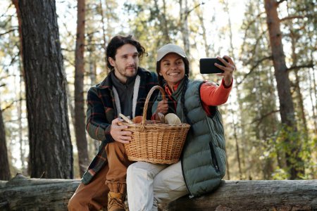 Téléchargez les photos : Deux jeunes cueilleurs de champignons heureux en tenue décontractée prenant selfie sur smartphone tout en étant assis sur tronc d'arbre sec dans la forêt d'automne - en image libre de droit