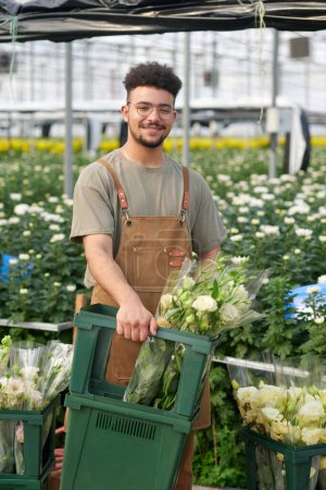 Téléchargez les photos : Heureux jeune travailleur masculin de serre industrielle avec des grappes de fleurs d'eustomie fraîches dans une boîte debout devant la caméra et vous regardant - en image libre de droit