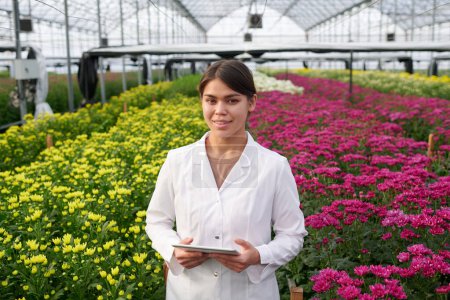 Foto de Joven agronomía femenina sonriente experta en labcoat usando tableta y mirando a la cámara mientras está de pie entre dos macizos de flores largos y anchos - Imagen libre de derechos