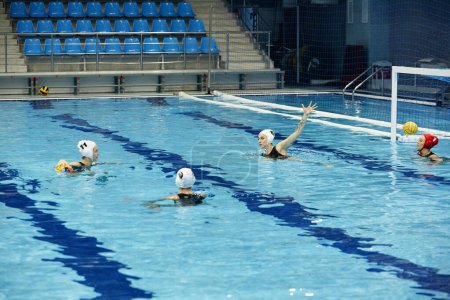 Téléchargez les photos : Deux joueurs de water-polo attaquent l'équipe rivale tandis que l'une des filles lance le ballon à la porte avec la sportive de défense pendant le match dans la piscine - en image libre de droit
