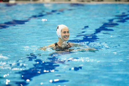 Téléchargez les photos : Tête de jeune femme souriante en bonnet de bain debout dans l'eau bleue profonde de la piscine et en regardant l'un des joueurs de water-polo pendant le jeu - en image libre de droit