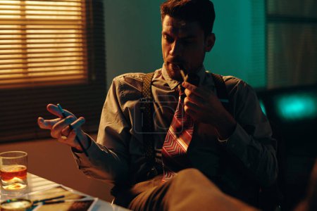 Téléchargez les photos : Jeune avocat ou policier pensif fumant dans un bureau sombre assis sur le lieu de travail et regardant à travers les photos sur la table - en image libre de droit