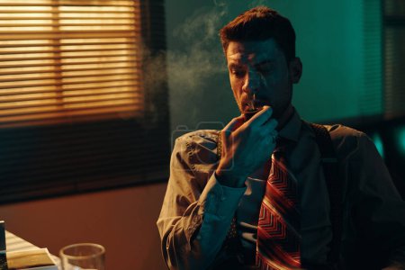 Téléchargez les photos : Jeune homme pensif avec pipe à tabac dans la bouche assis sur le lieu de travail devant la caméra pendant le travail dans un bureau sombre avec stores sur la fenêtre - en image libre de droit