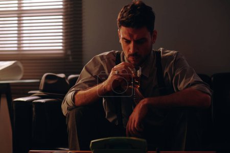 Téléchargez les photos : Homme d'affaires sérieux regardant du whisky dans un verre assis sur un canapé dans un bureau sombre avec une fenêtre couverte de stores vénitiens - en image libre de droit