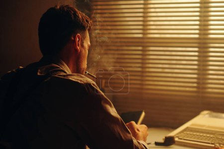 Foto de Vista trasera del joven fumando cigarrillo y mirando el libro en sus manos mientras está sentado frente a la ventana con persianas venecianas - Imagen libre de derechos