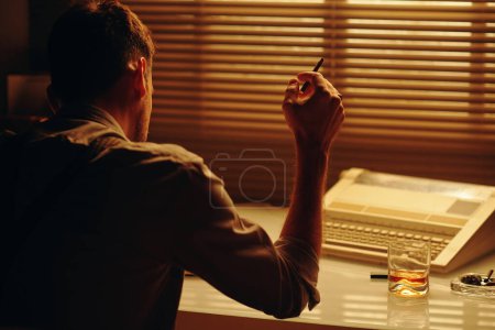 Photo for Rear view of young male clerk or police officer with cigarette in hand sitting by workplace in dark office and concentrating on work - Royalty Free Image