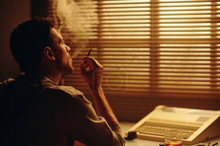Téléchargez les photos : Jeune homme inspiré fumant la cigarette et regardant par la fenêtre avec des stores vénitiens tout en étant assis près du lieu de travail avec une machine à écrire - en image libre de droit