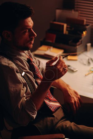 Foto de Vista lateral del joven hombre de negocios cansado con fumar cigarrillo en la mano sentado en el lugar de trabajo delante de la cámara y teniendo descanso - Imagen libre de derechos