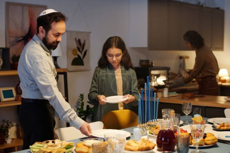 Téléchargez les photos : Homme mûr et sa fille mettre des assiettes et d'autres couverts sur la table du dîner avec de la nourriture juive traditionnelle maison préparée pour Hanoukka - en image libre de droit