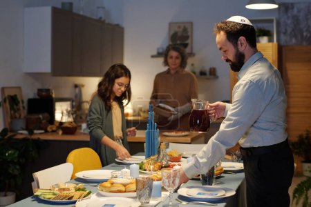 Téléchargez les photos : Vue latérale de l'homme barbu en chemise blanche aidant à servir la table avec des assiettes et une cruche en verre de jus fait maison pour la célébration de Hanoukka - en image libre de droit