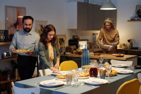 Foto de Linda chica judía ayudando a sus padres servir mesa mientras se pliega la servilleta en el plato contra su padre llevando bandeja con bocadillo fresco y plato - Imagen libre de derechos