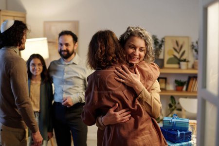 Photo for Mature cheerful woman embracing her daughter or guest while meeting and greeting her against other members of family at home - Royalty Free Image