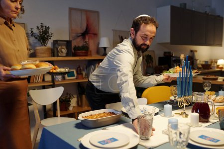 Foto de Judío barbudo en yarmulke ayudando a su esposa a servir mesa festiva con comida y bebidas caseras mientras espera a los invitados - Imagen libre de derechos