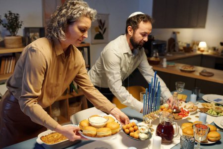 Foto de Mujer madura poniendo platos con rosquillas caseras y humus en la mesa que se sirve para los huéspedes antes de la celebración de Hanukkah - Imagen libre de derechos