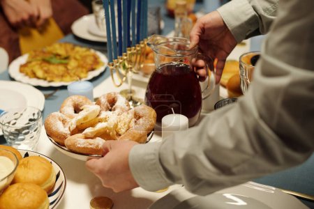 Téléchargez les photos : Mains d'homme juif avec cruche de jus de canneberge et beignets faits maison les mettre sur la table servi avec de la nourriture appétissante pour le dîner - en image libre de droit