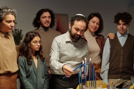 Foto de Hombre judío maduro en kippah encendiendo velas en el candelero de menorah mientras se inclina sobre la mesa servida entre los miembros de su familia - Imagen libre de derechos