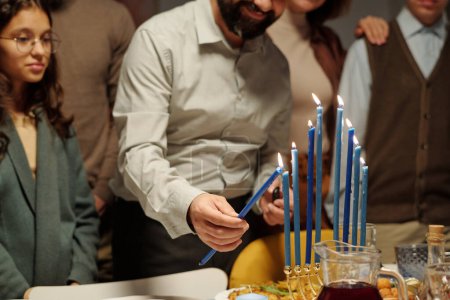 Téléchargez les photos : Homme barbu allumant l'une des bougies de menorah sur Hanoukka fête tout en se tenant à table servie parmi sa fille et d'autres membres de la famille - en image libre de droit