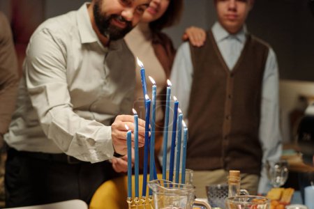Foto de Recortado tiro de hombre maduro poner novena vela en el candelero menorah de pie en la mesa servida con comida casera para Hanukkah - Imagen libre de derechos