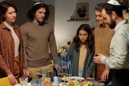 Foto de Linda chica en gafas encendiendo una de las velas menorah en la mesa servida mientras está de pie entre su familia y celebrando Hanukkah - Imagen libre de derechos