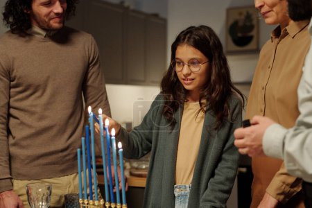 Photo for Pretty pre-teen girl lighting blue candles of menorah while standing among her family and preparing for Hanukkah celebration - Royalty Free Image