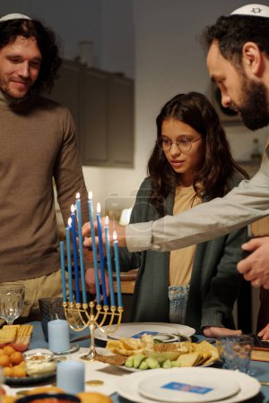 Foto de Hombre maduro ayudando a su hija a encender velas de menorah mientras se prepara para la celebración de Hanukkah por mesa servida - Imagen libre de derechos