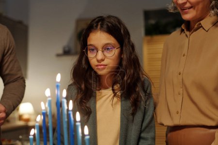 Téléchargez les photos : Adorable pré-adolescente dans des lunettes de vue regardant brûler des bougies bleues sur chandelier menorah tout en se tenant à côté de sa mère souriante - en image libre de droit