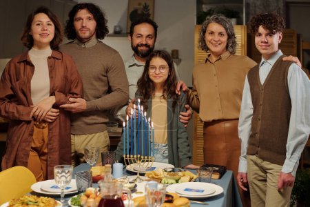 Téléchargez les photos : Famille de six membres de différentes générations debout à table servie avec des bougies menorah brûlantes et de la nourriture maison et vous regardant - en image libre de droit