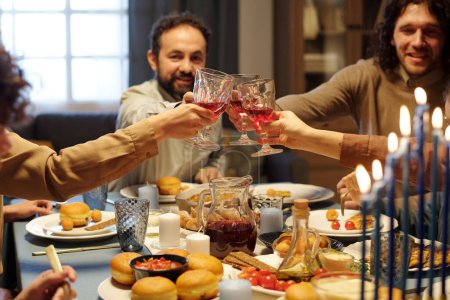 Téléchargez les photos : Les mains des membres de la famille juive cliquetis avec des verres de vin rouge sur la table de fête servi avec des collations faites maison pendant la célébration - en image libre de droit