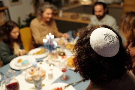 Foto de Yarmulke blanco con la estrella de plata de David en la cabeza del joven judío sentado junto a la mesa servida con comida y bebidas caseras - Imagen libre de derechos