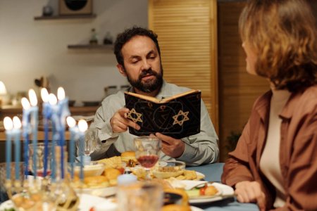 Téléchargez les photos : Homme juif mûr sérieux avec Torah ouverte lisant des textes de l'Écriture ou des Psaumes assis à table avec des membres de sa famille - en image libre de droit