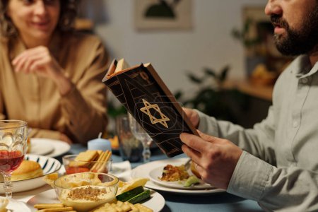 Téléchargez les photos : Homme juif barbu lisant le texte de la Torah à table pendant qu'il était assis devant les membres de sa famille pendant la célébration de Hanoukka - en image libre de droit