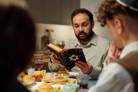 Foto de Enfócate en el rabino barbudo y el jefe de la familia judía leyendo el texto de la Torá mientras está sentado junto a la mesa servida frente a sus dos hijos. - Imagen libre de derechos