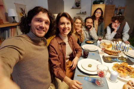 Téléchargez les photos : Joyeux jeune couple prenant selfie avec les membres de leur famille tout en étant assis près de la table de fête servie et profiter de Hanoukka dîner - en image libre de droit