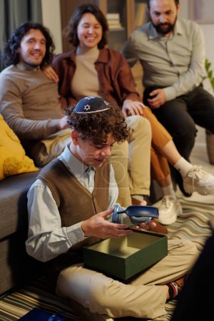 Téléchargez les photos : Pré-adolescent garçon juif avec casque déballer boîte cadeau avec Hanoukka cadeau de ses parents assis sur le sol par le canapé - en image libre de droit