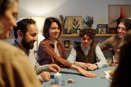 Téléchargez les photos : Jeune femme souriante mettre pièce sur le centre de la table tout en pariant pendant le jeu de loisirs avec les membres de sa famille après Hanoukka dîner - en image libre de droit