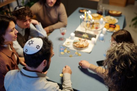 Téléchargez les photos : Angle élevé de l'homme juif mature avec kippah sur la tête assis parmi sa femme et sa fille pariant pendant le jeu de loisirs par table - en image libre de droit