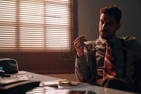 Foto de Joven hombre de negocios pensativo en ropa formal retro sosteniendo cigarrillo mientras está sentado junto a la mesa en la oficina oscura y pensando en algo - Imagen libre de derechos