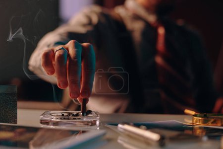 Téléchargez les photos : Close-up of hand of young man stubbing butt of cigarette in ashtray standing on table among business and office supplies - en image libre de droit