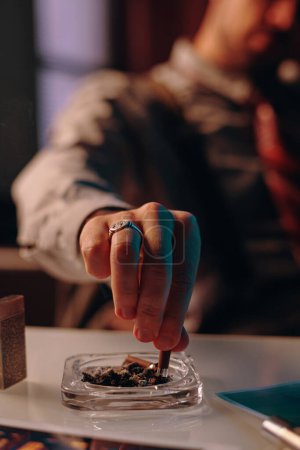 Photo for Hand of young man stubbing butt of cigarette in transparent square ashtray after smoking while sitting by table in office - Royalty Free Image
