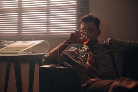 Téléchargez les photos : Jeune homme reposé en tenue de cérémonie prenant un verre de whisky assis sur un canapé en cuir noir contre une fenêtre avec stores vénitiens dans un bureau sombre - en image libre de droit