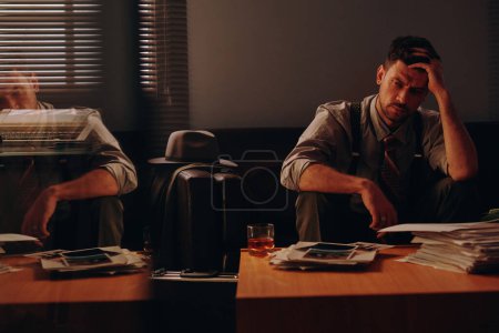 Téléchargez les photos : Jeune homme d'affaires surmené touchant la tête et regardant la pile de documents papier sur la table alors qu'il était assis sur le canapé dans un bureau sombre - en image libre de droit
