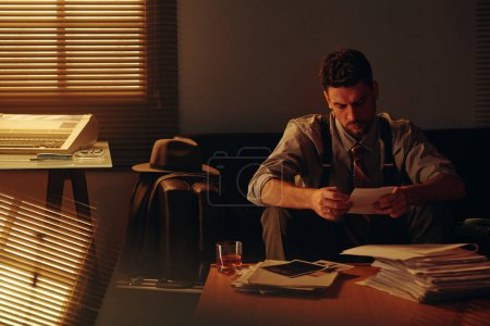 Foto de Joven empleado serio u oficial de policía mirando la foto mientras está sentado frente al lugar de trabajo con un montón de papeles y un vaso de whisky en la oficina - Imagen libre de derechos